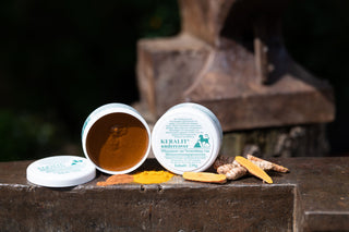 An open and a closed tin of Keralit Undercover care paste stand on an iron table with the ingredients turmeric and oak bark
