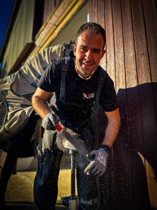 A man grooms a horse's hoof in front of a stable