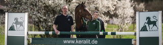 A man and a woman stand together with a brown horse behind an obstacle that readskeralit