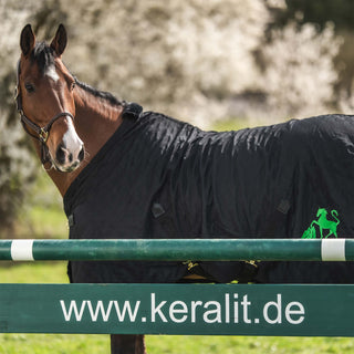 Ein braunes Pferd mit einer Decke steht hinter einem Hindernis mit der Aufschrift www.keralit.de