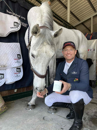 A man in dressage clothes kneels in front of a white horse in the stable and holds the Keralit laurel ointment in his hand